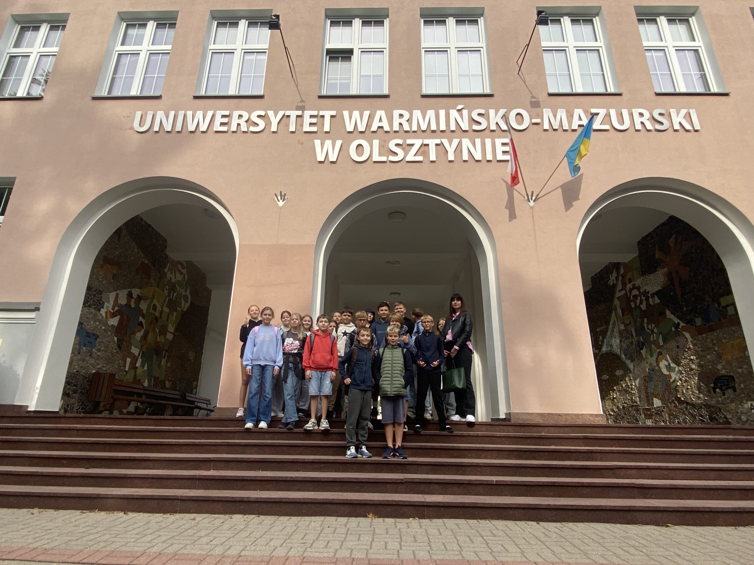 Na zdjęciu Grupka dzieci stoi na schodach przed Rektoratem uniwersytetu warmińsko-mazurskiego. Stoją one pod napisem uniwersytet warmińsko mazurski w Olsztynie. Dzieci stoją na tle budynku który posiada trzy wejścia w kształcie łuku. Budynek jest koloru rudawego na budynku wiszą dwie flagi, Jedna jest to flaga biało czerwona druga niebieska flaga uniwersytetu warmińsko-mazurskiego.