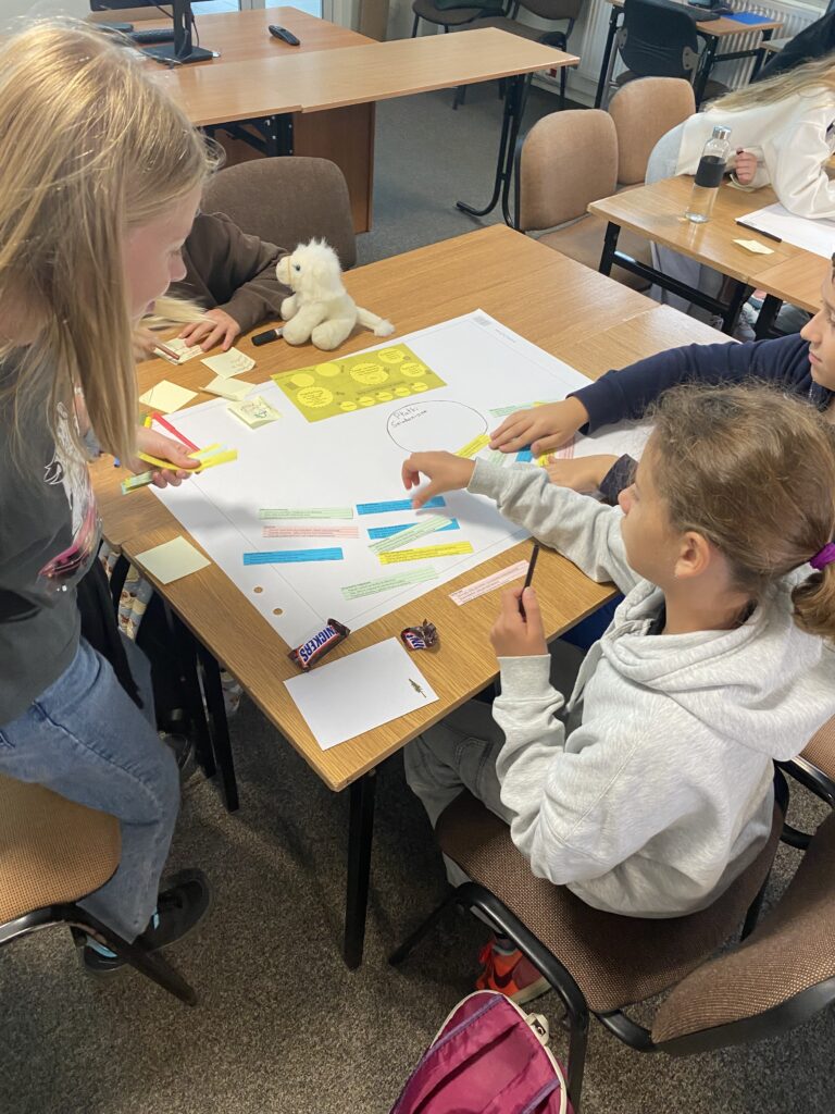 Na zdjęciu widać dwie dziewczynki i trochę jednego chłopca którzy siedzą przy brązowym stoliku. Na stoliku leży duży Arkusz białego papieru na tym arkuszu rozłożone są kolorowe małe Karteczki pocięte w długie prostokąty. Z lewej strony stolika stoi dziewczynka w Blond włosach pochyla się nad pracą. Obok siedzi dziewczynka w białej bluzie i ciemnych włoskach na krześle i ręką chwyta karteczkę o kolorze niebieskim. Chłopiec siedzący obok jest w ciemnej bluzie i ręką przykrywa dwa paseczki koloru żółtego i niebieskiego. Na podłodze stoi różowy plecak. Na ławce widać też plucia czka białego małego przypominającego konia. W tle widać ławki i biurka.