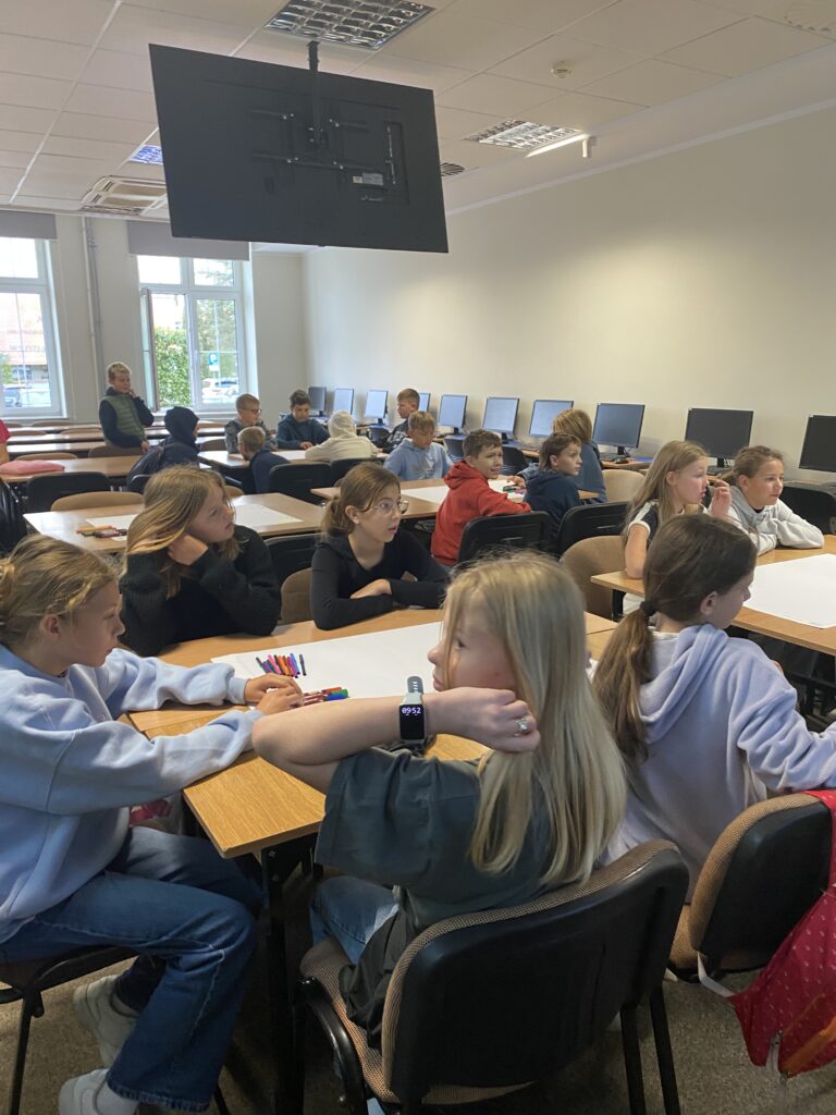 Grupa dzieci siedzi w ławkach w sali ćwiczeniowej, w której są komputer dookoła ściany. ławki są brązowe, Ściany są pomalowane na jasny kolor. dzieci siedzą zwrócone w stronę projektora którego nie widać na zdjęciu. Nad dziećmi wisi ekran na którym wyświetlane są informacje ekran ten na zdjęciu jest tyle więc nie widać co na nim jest.