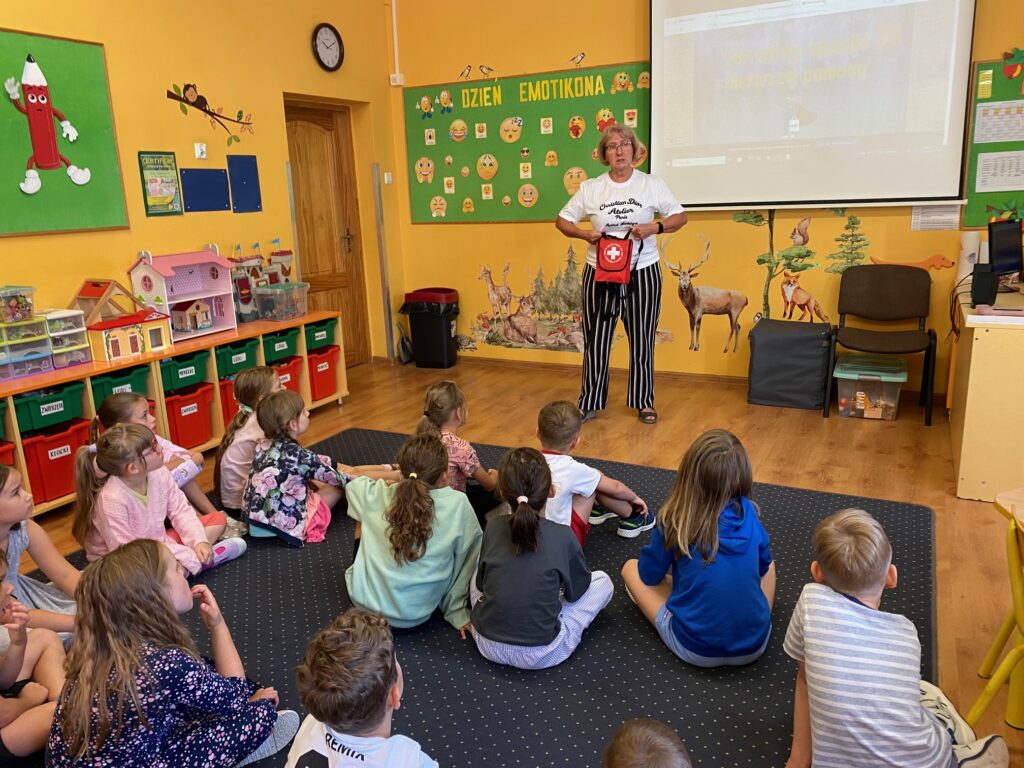 Zdjęcie przedstawia siedzące w świetlicy dzieci. Dzieci siedzą tyłem, ubrane są w kolorowe ubrania. Patrzą na stojącą przodem panią pielęgniarkę. Trzyma ona w rękach czerwoną torbę - apteczkę pierwszej pomocy. Ściany świetlicy są koloru żółtego. Wiszą na nich kolorowe gazetki ścienne a na podłodze stoją szafki z kolorowymi zabawkami.