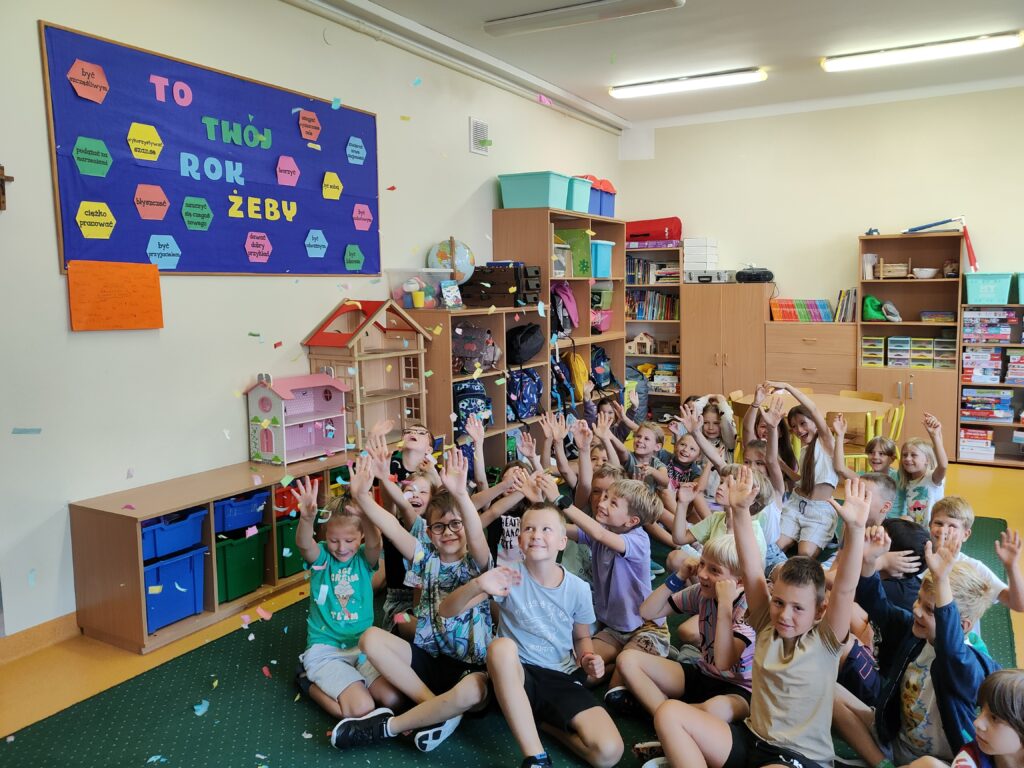 W sali świetlicy na zielonym dywanie siedzi grupa kolorowo ubranych dzieci. Ręce dzieci są podniesione do góry. W powietrzu unosi się kolorowe confetti. Lezy ono także na zielonym dywanie. Na ścianie koloru kremowego wisi kolorowa gazetka. Na drugim planie widać brązowe szafki z kolorowym zabawkami i plecakami.