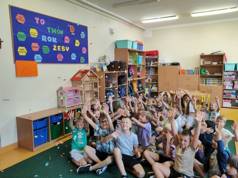 W sali świetlicy na zielonym dywanie siedzi grupa kolorowo ubranych dzieci. Ręce dzieci są podniesione do góry. W powietrzu unosi się kolorowe confetti. Lezy ono także na zielonym dywanie. Na ścianie koloru kremowego wisi kolorowa gazetka. Na drugim planie widać brązowe szafki z kolorowym zabawkami i plecakami.