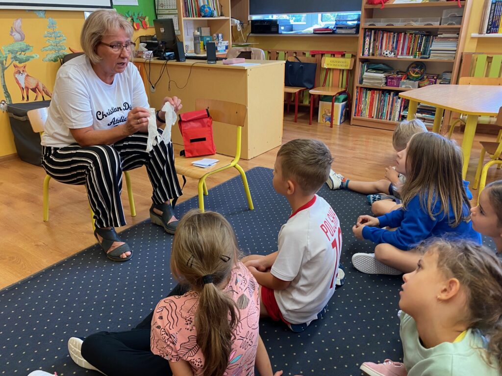 Zdjęcie przedstawia siedzące w świetlicy dzieci. Dzieci siedzą bokiem, ubrane są w kolorowe ubrania. Patrzą na siedzącą na żółtym krzesełku panią pielęgniarkę. Trzyma ona w rękach jednorazowe rękawiczki. Na krzesełku obok leży czerwona apteczka pierwszej pomocy. Ściany świetlicy są koloru żółtego. Na drugim planie stoi biurko, widać parapet okna.