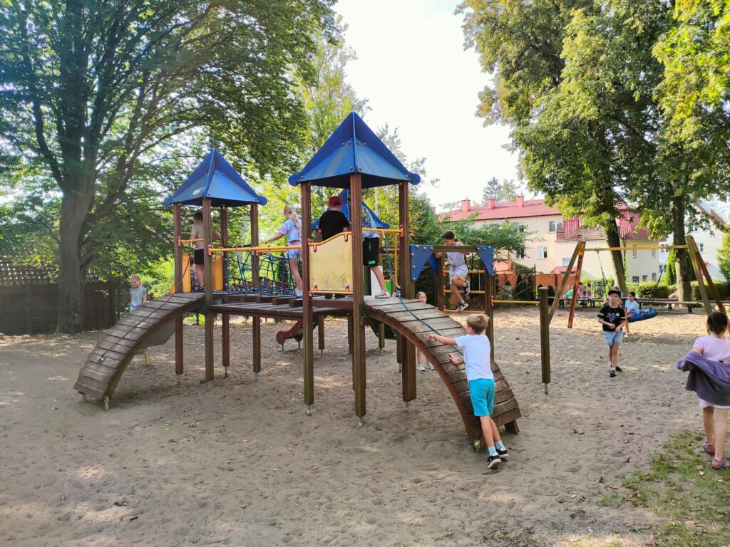 Zdjęcie to przedstawia dzieci bawiące się na placu zabaw. Na pierwszym planie widać drewniana konstrukcję „ domku”. Ma on dwie wieżyczki zakończone niebieskim daszkiem. Chłopiec w niebieskim ubraniu wspina się po drewnianej pochylni domku. Na placu zabaw piasek. Dalszy plan to bawiące się dzieci. W oddali zielone drzewa i dachy domów.