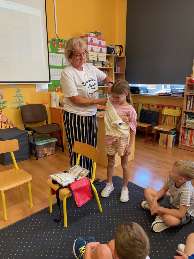 Zdjęcie przedstawia świetlicę. Dwoje dzieci siedzi bokiem na szarym dywanie, ubrane są w kolorowe ubrania. Patrzą na stojącą panią pielęgniarkę. Pielęgniarka zakłada temblak stojącej obok niej dziewczynce. Na żółtym krześle stojącym obok leży czerwona apteczka pierwszej pomocy. Ściany świetlicy są koloru żółtego. Na drugim planie stoi biurko, widać parapet okna i zasłonę okna koloru szarego.