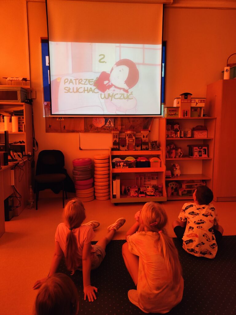 Zdjęcie przedstawia świetlicę. Zgaszone światło powoduje iż całe zdjęcie jest w odcieniach pomarańczowych. Na pierwszym planie czwórka dzieci siedzi tyłem na brązowym dywanie. Patrzą na projektor na którym wyświetlany jest film na temat pierwszej pomocy. Na dalszym planie oparte o ścianę brązowe krzesło i szafki z zabawkami.