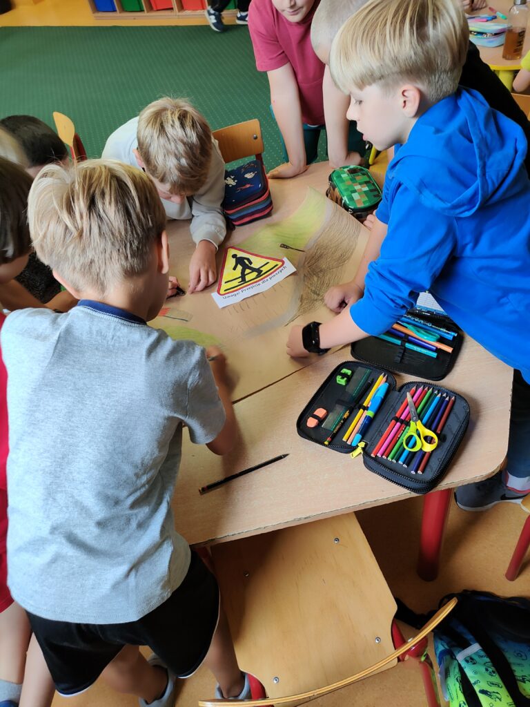 Zdjęcie ukazuje grupę chłopców, którzy stoją wokół stołu i tworzą pracę plastyczną. Chłopcy ubrani są w kolorowe ubrania a stół jest koloru brązowego. Na stole leży brązowy duży papier pakowy, na którym naklejona jest kolorowanka , przedstawiająca ostrzegawczy znak drogowy Przejście dla pieszych. Na drugim planie widać zielony dywan i kolorowe szafki.