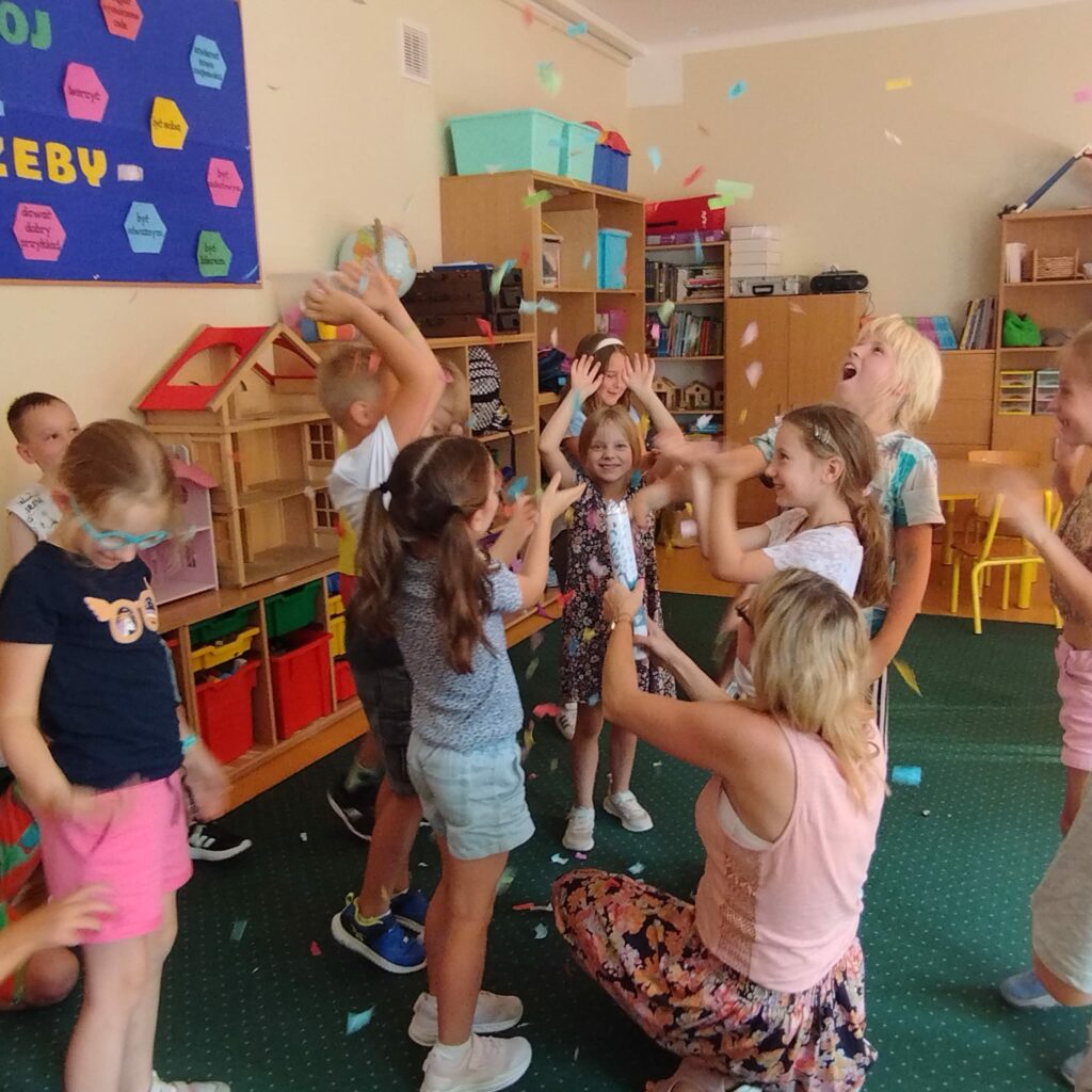 W sali świetlicy na zielonym dywanie stoi grupa kolorowo ubranych dzieci. Ręce dzieci są podniesione do góry. W powietrzu unosi się kolorowe confetti. Lezy ono także na zielonym dywanie. Na twarzach dzieci gości uśmiech. Na ścianie koloru kremowego wisi kolorowa gazetka. Na drugim planie widać brązowe szafki z kolorowym zabawkami i plecakami.