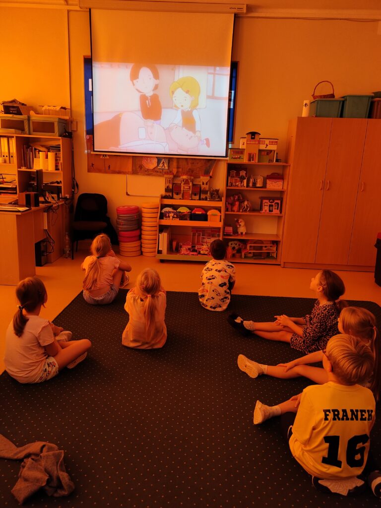 Zdjęcie przedstawia świetlicę. Zgaszone światło powoduje iż całe zdjęcie jest w odcieniach pomarańczowych. Na pierwszym planie siódemka dzieci siedzi tyłem na brązowym dywanie. Patrzą na projektor na którym wyświetlany jest film na temat pierwszej pomocy. Na dalszym planie oparte o ścianę brązowe krzesło i szafki z zabawkami.