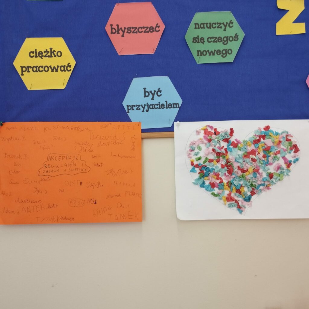 Zdjęcie przedstawia kolorowe prace plastyczne. Górna część zdjęcia to na niebieskim tle napisy: „ciężko pracować:, „błyszczeć”, „być przyjacielem”, „ nauczyć się czegoś nowego”. Dolna część zdjęcia to dwie prace plastyczne powieszone na kremowym tle. Pierwsza to pomarańczowa kartka z podpisami uczniów . Podpisywali , że akceptują zasady obowiązujące w świetlicy. Druga praca przedstawia kolorowe serce na białym tle. Serce wyklejone jest kolorowymi confetti.