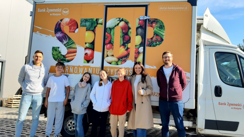 Na zdjęciu widać grupę siedmiu osób stojących przed ciężarówką z napisem „STOP marnowaniu żywności” oraz logo „Bank Żywności w Olsztynie”. Ciężarówka jest ozdobiona obrazkami przedstawiającymi różne warzywa i owoce. Wszyscy na zdjęciu uśmiechają się, niektórzy w wygodnych, codziennych ubraniach, a inni w kurtkach lub bluzach. Tło wskazuje, że zdjęcie zostało zrobione na zewnątrz, prawdopodobnie podczas wydarzenia lub akcji związanej z bankiem żywności, mającej na celu walkę z marnowaniem jedzenia.