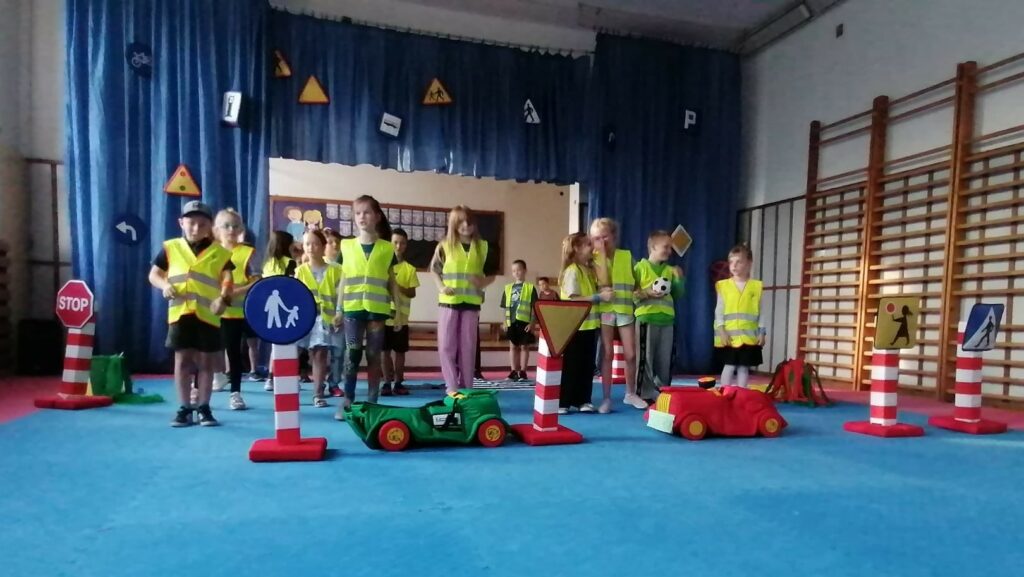 Na sali gimnastycznej stoi grupa dzieci. Ubrane są w kamizelki odblaskowe i śpiewają piosenkę. Za nimi znajduje się scena ozdobiona niebieskim materiałem. Na nim przyczepione są różne znaki drogowe. Przed nimi i z boku, jest dekoracja – znaki drogowe, umieszczone na czerwono- białych podstawach oraz 2 samochody, uszyte z materiału.