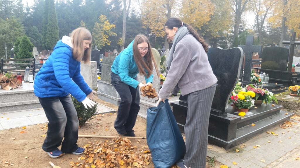 Na zdjęciu widać trzy młode osoby pracujące na cmentarzu. Są ubrane w ciepłe kurtki, co sugeruje, że jest chłodna pora roku. Wszystkie osoby mają na rękach rękawiczki, a jedna z nich zbiera liście i wrzuca je do dużego, niebieskiego worka, który trzyma inna osoba. W tle widoczne są nagrobki oraz ozdoby kwiatowe, co potwierdza, że akcja ma miejsce na cmentarzu. Drzewa w tle mają żółte liście, co może wskazywać na jesień. Cała scena przedstawia zaangażowanie i współpracę w porządkowaniu tego miejsca.