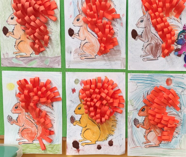 zdjęcie przedstawia 6 prac plastycznych z postacią wiewiórki, która trzyma w łapce brązowego orzeszka i jest pokolorowana pomarańczową kredką. Ogon wyklejony jest pomarańczowymi paskami papieru sklejonymi końcami. Tak wykonany ogon daje efekt przestrzenności pracy plastycznej.