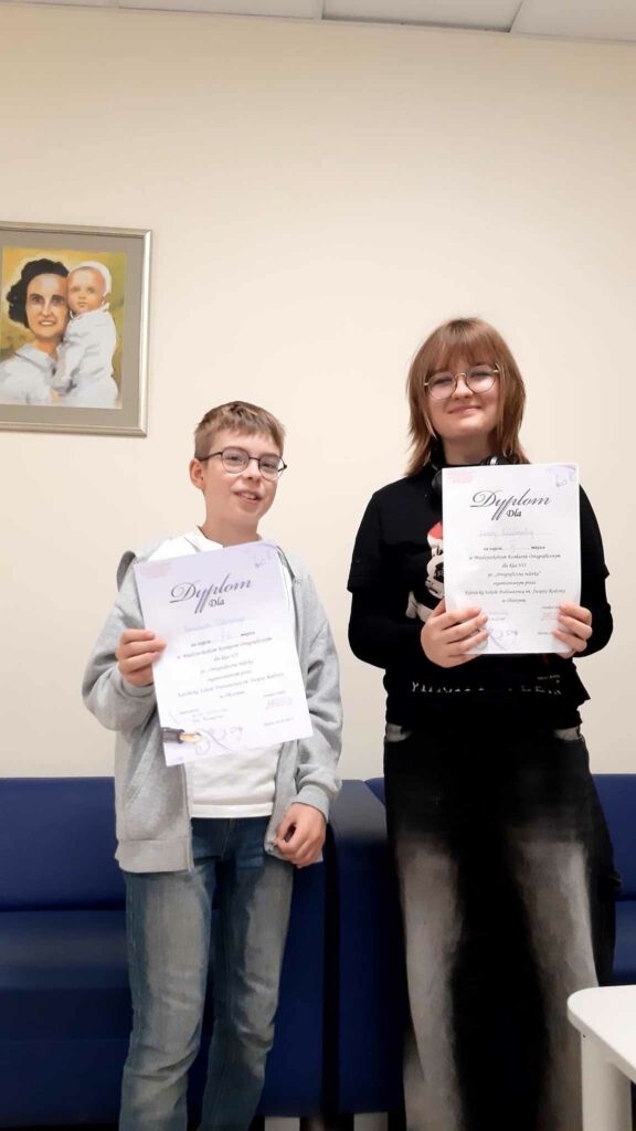 Dwoje uczniów dziewczyna i chłopak stoją obok siebie, trzymając w dłoniach dyplomy. Na ich twarzach widać uśmiechy, które świadczą o zadowoleniu z osiągnięcia zajętego miejsca w międzyszkolnym konkursie ortograficznym. W tle na ścianie wisi obraz, który dodaje formalnego charakteru temu uroczystemu momentowi.