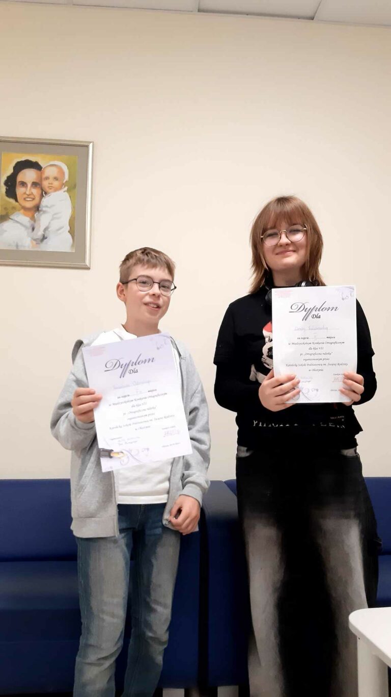 Dwoje uczniów dziewczyna i chłopak stoją obok siebie, trzymając w dłoniach dyplomy. Na ich twarzach widać uśmiechy, które świadczą o zadowoleniu z osiągnięcia zajętego miejsca w międzyszkolnym konkursie ortograficznym. W tle na ścianie wisi obraz, który dodaje formalnego charakteru temu uroczystemu momentowi.