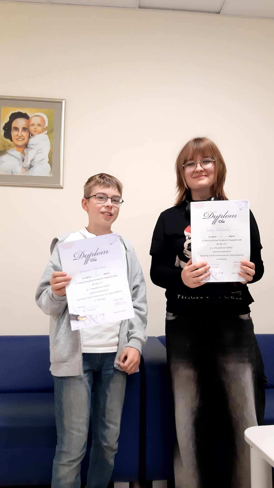 Dwoje uczniów dziewczyna i chłopak stoją obok siebie, trzymając w dłoniach dyplomy. Na ich twarzach widać uśmiechy, które świadczą o zadowoleniu z osiągnięcia zajętego miejsca w międzyszkolnym konkursie ortograficznym. W tle na ścianie wisi obraz, który dodaje formalnego charakteru temu uroczystemu momentowi.