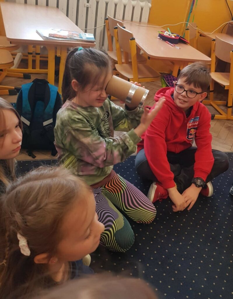 Na zdjęciu widzimy grupę dzieci siedzących w kręgu na dywanie w sali szkolnej. Dziewczynka w centrum trzyma przedmiot przypominający teleskop zrobiony z kartonowej tuby, przez który próbuje coś zobaczyć. Obok niej chłopiec w czerwonej bluzie, z okularami na nosie, patrzy na nią z zaciekawieniem. W tle widać szkolne biurka i krzesła oraz kolorowe przybory do rysowania na stole.