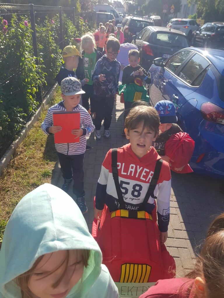 Widać grupę dzieci, idących chodnikiem. Na I planie widać dziewczynkę z kapturem na głowie. Za nią idzie chłopie ubrany w miękki samochód na szelkach. Dalej jeszcze kilkanaścioro dzieci idzie i niesie elementy dekoracji z apelu. Dzieci zajmują cały chodnik, obok stoją zaparkowane samochody.