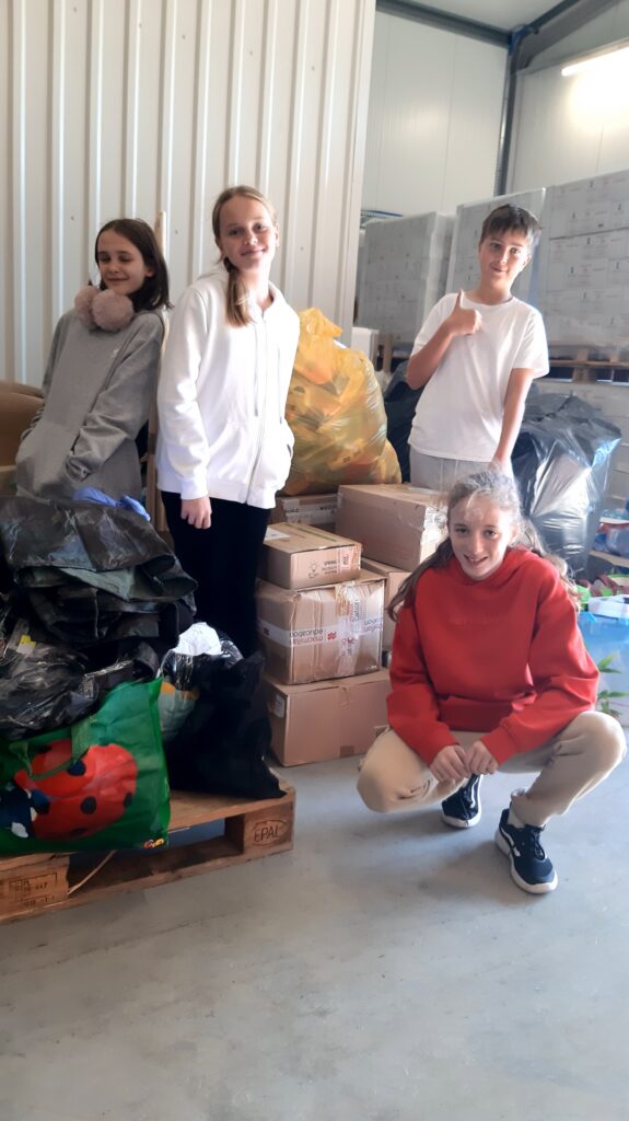 Na zdjęciu znajduje się czworo młodych ludzi, którzy stoją i kucają obok stosu paczek i worków. Zdjęcie wykonano wewnątrz pomieszczenia o jasnych ścianach z metalowymi panelami, co sugeruje, że jest to magazyn lub hala. W tle widoczne są zapakowane kartony oraz worki, co może sugerować, że młodzież bierze udział w akcji sortowania lub dystrybucji żywności lub innych produktów. Na pierwszym planie widać paczki, kartony i worki, ułożone na drewnianej palecie, co może świadczyć o trwającej pracy związanej z rozładunkiem lub przygotowaniem paczek do dalszej dystrybucji w ramach jakiejś charytatywnej akcji.