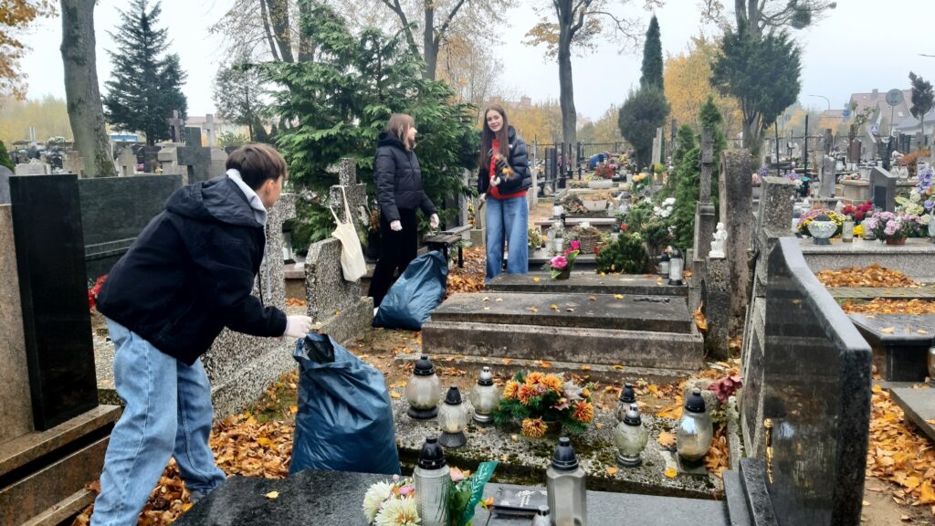 Na zdjęciu widzimy grupę młodych osób biorących udział w akcji sprzątania cmentarza. Troje wolontariuszy jest zajętych zbieraniem liści, które wkładają do dużych, niebieskich worków na śmieci. Wszystkie osoby są ubrane w ciepłe, zimowe kurtki, co sugeruje chłodną, jesienną pogodę. W tle widoczne są nagrobki ozdobione kwiatami i zniczami, a alejki są pokryte opadłymi liśćmi, co podkreśla jesienną atmosferę. Widać, że praca przebiega w miłej atmosferze, a uczestnicy chętnie angażują się w utrzymanie porządku na cmentarzu