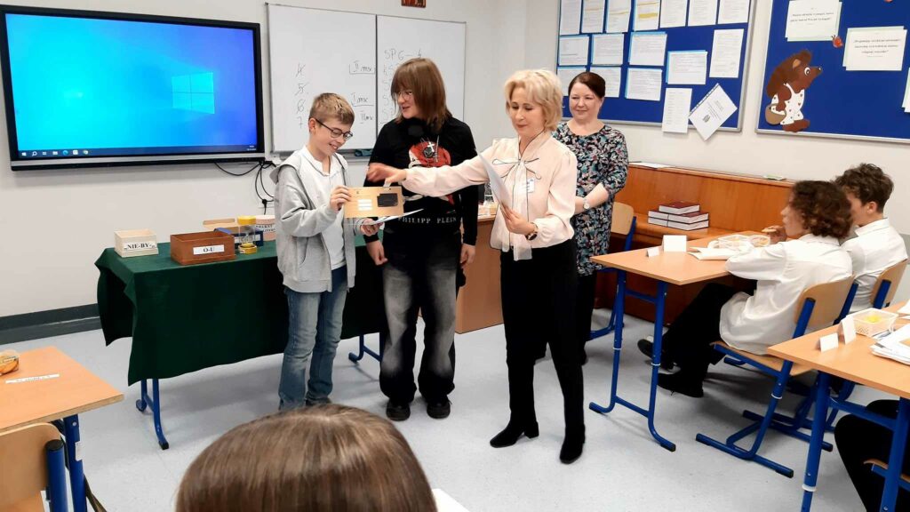 Na zdjęciu widzimy moment wręczania nagród. Uczniowie z klasy 7a, stojąc obok siebie, odbierają gratulacje od organizatorki konkursu. W tle znajduje się tablica z wyświetlonym ekranem, a na stole stoją przygotowane materiały do konkursu. Obok uczniów stoją także dwie osoby dorosłe, prawdopodobnie nauczycielki lub członkinie jury.