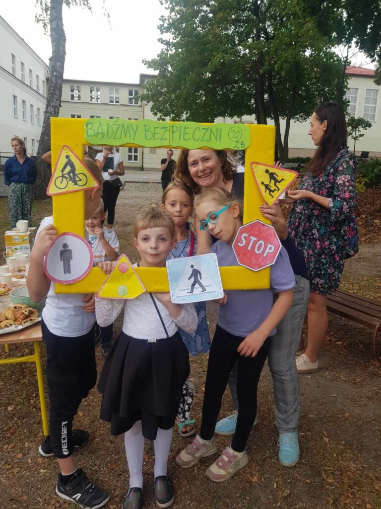 Na boisku szkolnym, na i planie widać kilkoro dzieci i nauczycielkę. Pozują oni w tzw. foto ramce z napisem Bądźmy bezpieczni i znakami drogowymi. W ramce widać głowy tych osób, poza nią jest reszta sylwetek. Po lewej stronie widać fragment stołu z poczęstunkiem.