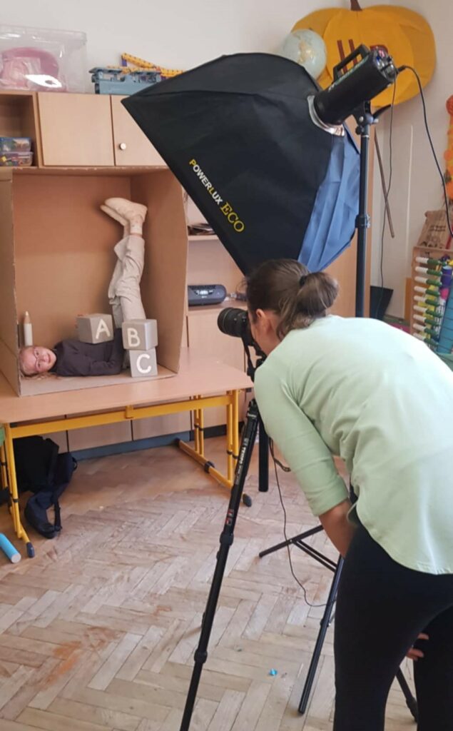 Na zdjęciu widać sesję fotograficzną w szkolnej klasie. Fotografka robi zdjęcie dziecka leżącego w dużym kartonowym pudle, ustawionego w zabawnej pozie, z nogami uniesionymi do góry. Dziecko otoczone jest drewnianymi klockami z literami "A", "B" i "C", a także dużą kredką. W tle widać sprzęt do oświetlenia, zabawki oraz inne elementy sali lekcyjnej.