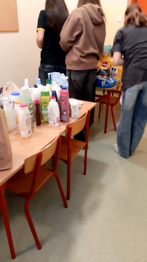 Na zdjęciu widać grupę osób, które stoją przy stole w pomieszczeniu. Osoby są odwrócone tyłem do kamery, zajęte układaniem lub sortowaniem różnych produktów. Na pierwszym planie znajduje się stół, na którym ustawione są różnego rodzaju butelki i opakowania – są to głównie produkty higieniczne, takie jak szampony, żele pod prysznic, balsamy i mydła. Produkty są różnokolorowe, a ich ustawienie sugeruje, że są przygotowywane do dalszego użytku lub pakowania.