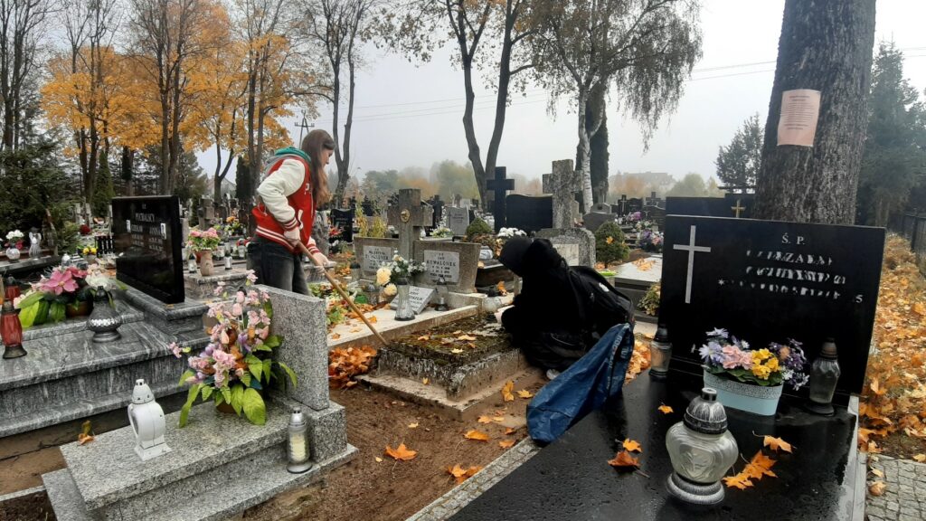 Na zdjęciu widzimy dwie osoby, które uczestniczą w sprzątaniu cmentarza. Jedna z nich, ubrana w czerwoną kurtkę z białymi rękawami, pracuje przy użyciu grabi, uprzątając liście z grobu. Druga osoba, ubrana na czarno, kuca przy jednym z nagrobków, prawdopodobnie oczyszczając go ręcznie. W tle widoczne są liczne nagrobki ozdobione kwiatami, zniczami i innymi dekoracjami, co podkreśla atmosferę troski o zmarłych. Jesienna sceneria, z drzewami o żółtych liściach i mglistym niebem, dodaje nastrojowego klimatu całemu wydarzeniu. Wolontariusze wydają się być zaangażowani w swoją pracę, porządkując groby z należytą starannością.