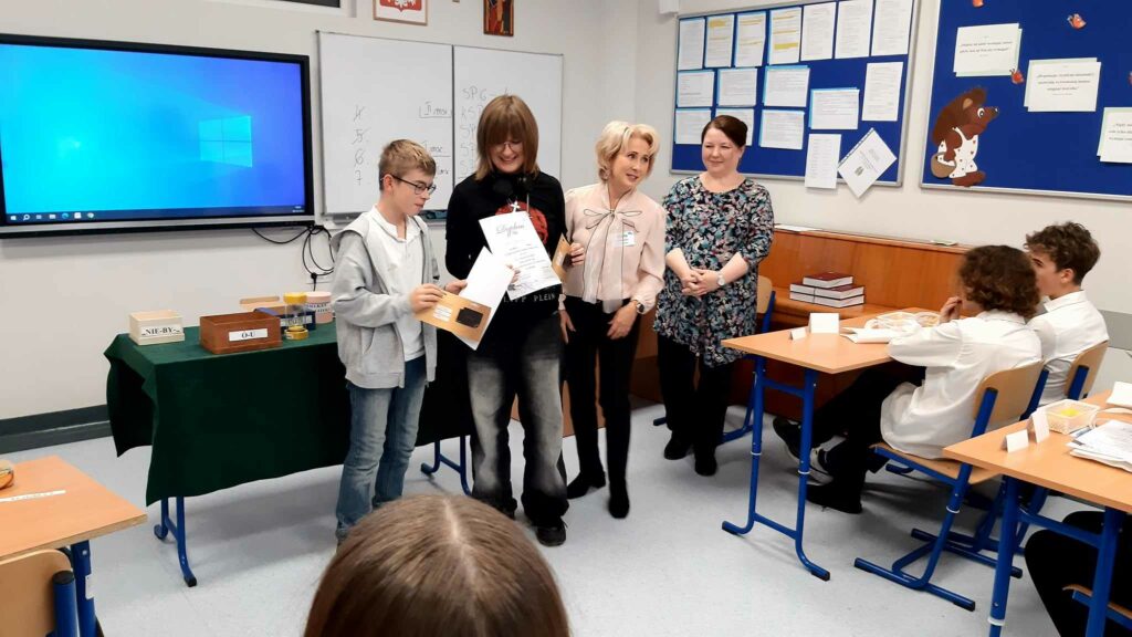 To kolejna scena wręczenia nagród. Uczniowie trzymają dyplomy i stoją obok organizatorki konkursu, która uśmiecha się i gratuluje im sukcesu. W tle widać siedzących przy ławkach innych uczestników, którzy przyglądają się ceremonii zakończenia konkursu.