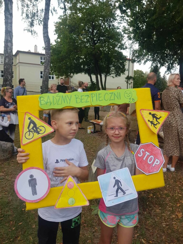 Na terenie przyszkolnym widać chłopca i dziewczynkę. Dzieci trzymają foto ramkę, ozdobioną znakami drogowymi i napisem Bądźmy bezpieczni. Dziewczynka nosi okulary, jest uśmiechnięta, chłopiec patrzy w bok..Za nimi, w tle widać rodziców.