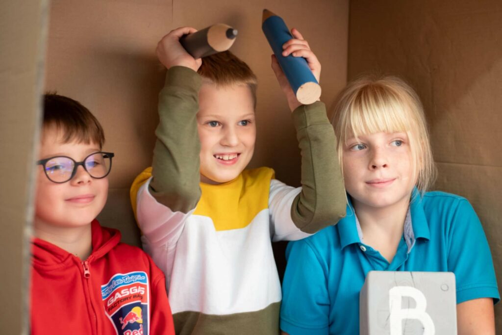 Na zdjęciu widać sesję zdjęciową w klasie. trzech uśmiechniętych chłopców pozuje do zdjecia. środkowy chłopiec trzyma w górze rekwizyty- 2 duże kredki, trzeci chłopec trzba klocek z literą B. Chłopcy pozują na tle kartonu.