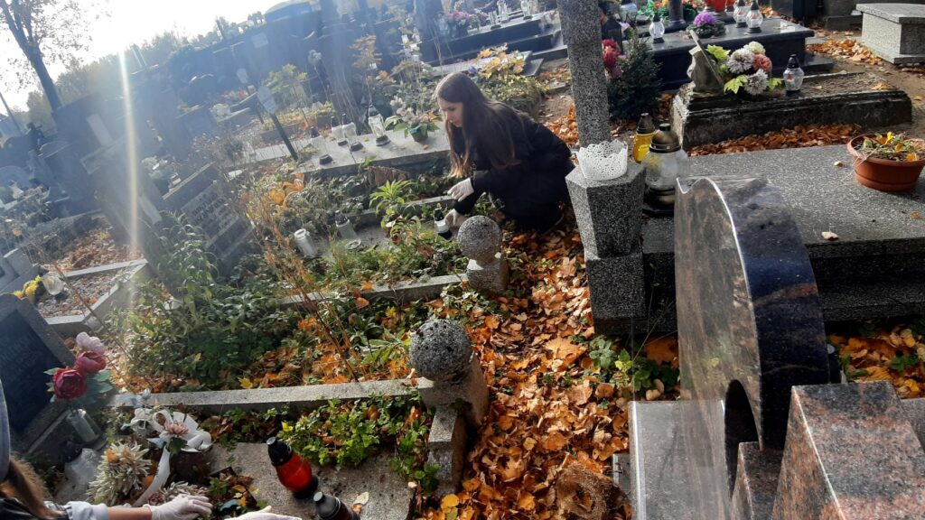 Na zdjęciu widać dziewczynę klęczącą przy grobie, trzymającą w dłoniach znicz. Maja na sobie rękawiczki, w celu ochrony dłoni, jest otoczona opadłymi jesiennymi liśćmi, które pokrywają cmentarz. Grób jest ozdobiony roślinami, kwiatami i świecami, które są typowymi elementami tego typu otoczenia. W tle widać inne nagrobki, wiele z nich również ozdobionych kwiatami i świecami. Słońce świeci jasno, rzucając ukośny promień na obraz, dodając scenie spokojnej i cichej atmosfery. Osoba wydaje się skupiona na swoim zadaniu, przyczyniając się do opieki i utrzymania grobu.