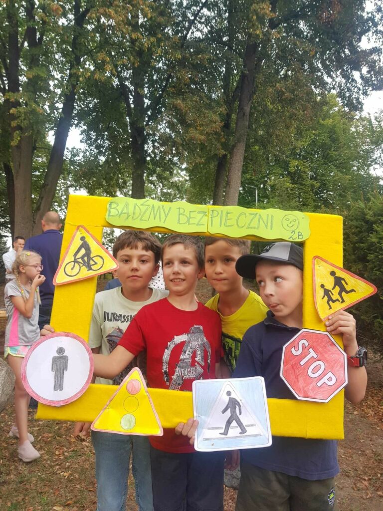 Na terenie przyszkolnym, na tle drzew, widać czterech chłopców. Dzieci trzymają foto ramkę, ozdobioną znakami drogowymi i napisem Bądźmy bezpieczni. Obok widać idąca dziewczynkę.