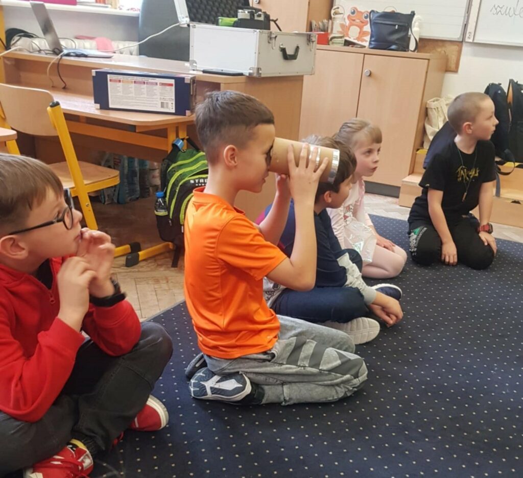 Na zdjęciu widzimy grupę dzieci siedzących na dywanie w sali szkolnej. Chłopiec w centrum trzyma przedmiot przypominający teleskop zrobiony z kartonowej tuby, przez który próbuje coś zobaczyć. W tle widać szkolne biurka i krzesła oraz kolorowe przybory do rysowania na stole.