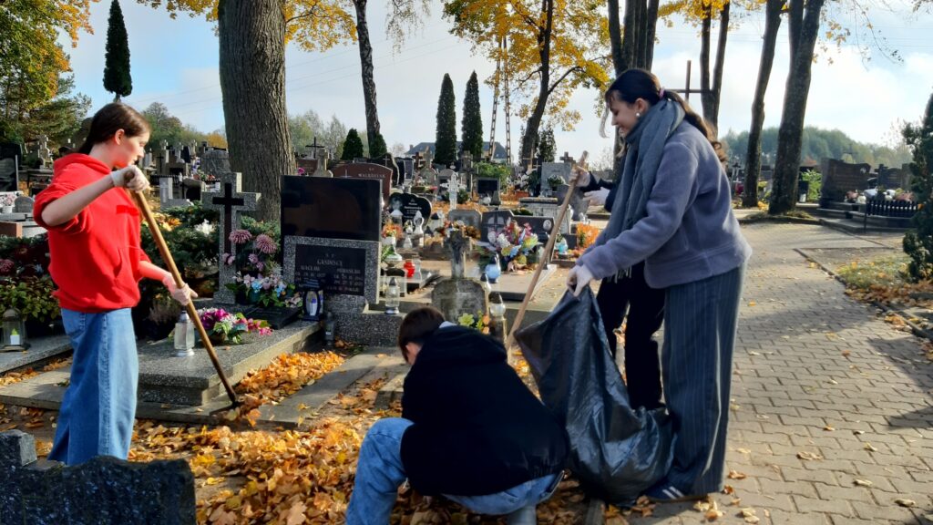 Na zdjęciu widać trzy osoby pracujące razem na cmentarzu, sprzątające opadłe liście. Jedna osoba, ubrana w czerwoną bluzę z kapturem, używa grabi do zbierania liści, podczas gdy druga osoba, w jasnofioletowej kurtce i szaliku, trzyma otwarty duży worek na śmieci. Trzecia osoba kuca, prawdopodobnie zbierając liście lub inne śmieci, aby umieścić je w worku. Nagrobki w tle są ozdobione kwiatami, świecami i roślinami, co podkreśla dbałość o utrzymanie grobów. Atmosfera jest spokojna, a światło słoneczne prześwieca przez jesienne liście, tworząc ciepłe i spokojne otoczenie. Scena odzwierciedla wspólny wysiłek, aby utrzymać cmentarz w czystości i szacunku.