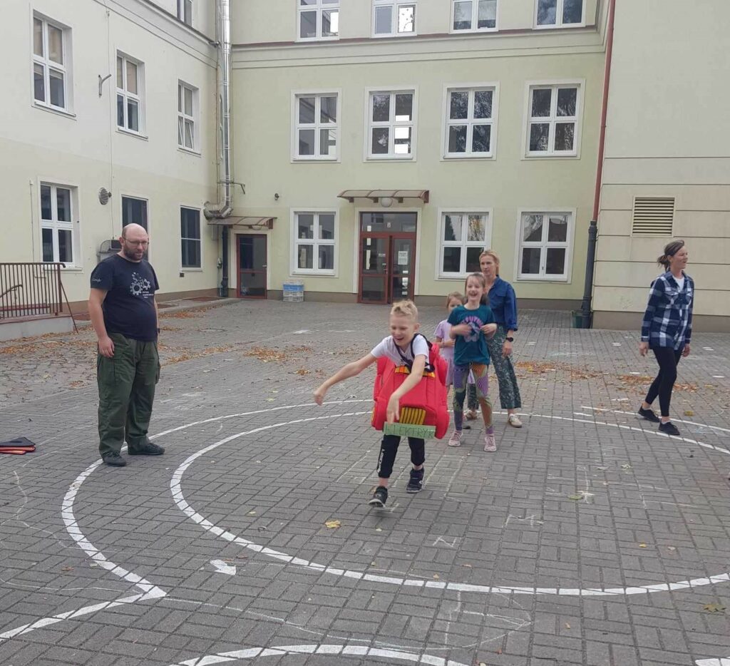 Na zdjęciu widać wybrukowany teren przed szkołą. Na nim wymalowane są białe linie. Na I planie widać biegnącego chłopca. Chłopiec ma założony na sobie tzw.autochodzik (miękkie auto na szelkach). Za nim czekają na swoją kolej 2 dziewczynki. Po jednej i drugiej stronie stoi dwójka rodziców: patrzą i dopingują.