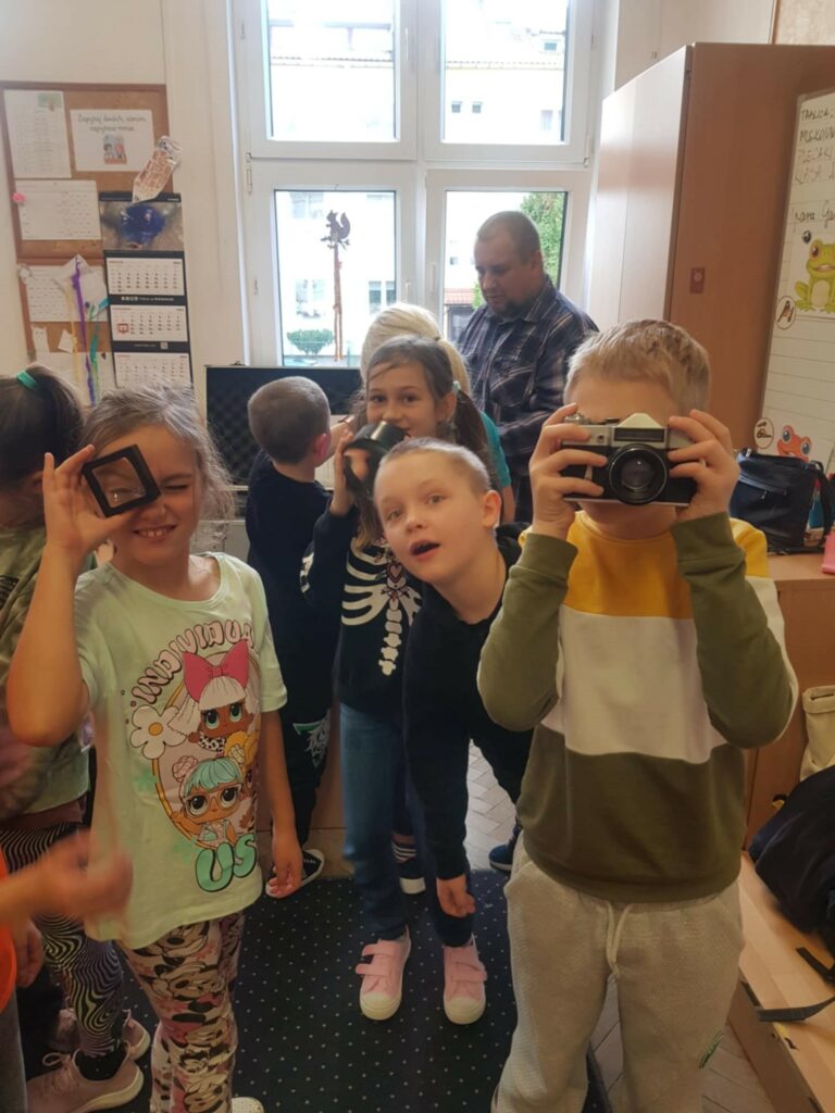 Kilkoro dzieci stoi w klasie na tle okna. Troje z nich trzyma przedmioty związane z fotografią: lupę, aparat. Za nimi stoi mężczyzna, który pochyla się nad walizką ze sprzętem.