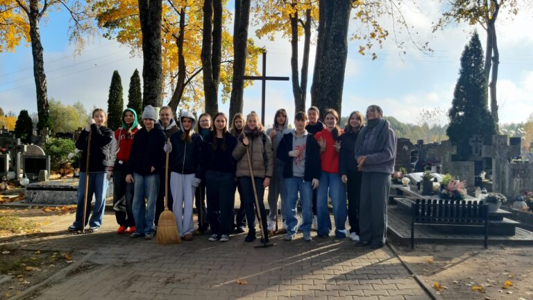 Na zdjęciu widać grupę ludzi stojących razem na cmentarzu, otoczonych nagrobkami i drzewami z jesiennymi liśćmi. Grupa składa się z około 14 osób, głównie młodych ludzi, niektórzy trzymają narzędzia, takie jak miotły i grabie, co sugeruje, że mogą być zajęci sprzątaniem lub konserwacją cmentarza. Pogoda wydaje się być chłodna, ponieważ niektórzy noszą kurtki, czapki i szaliki. W tle widać duży krzyż i nagrobki ozdobione kwiatami, co wskazuje, że jest to rzeczywiście miejsce na cmentarzu. Ogólna atmosfera jest spokojna i pełna szacunku, z poczuciem wysiłku społeczności.