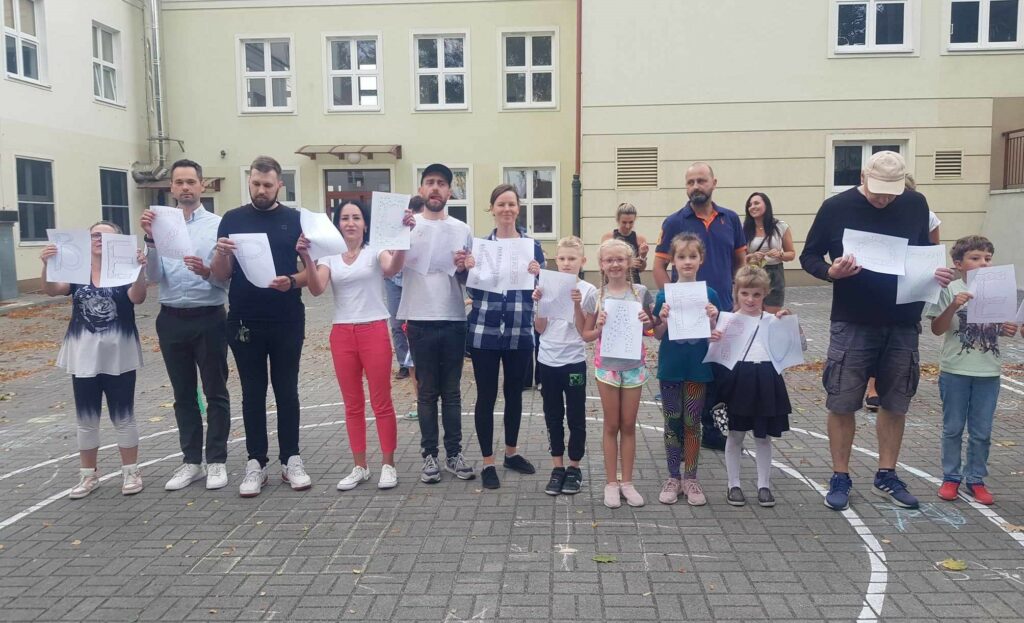 Na terenie, przed szkołą, stoją w szeregu rodzice z dziećmi. Każdy trzyma w ręku podniesioną kartkę. Z liter powstało hasło Bezpieczni na drodze.