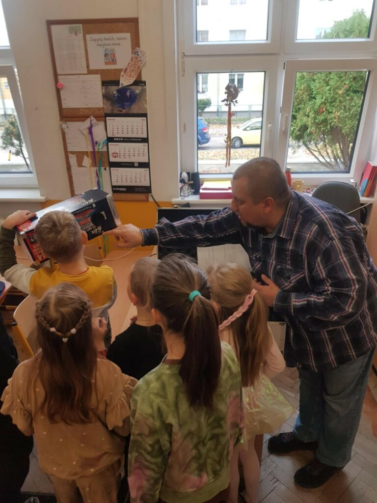 na zdjęciu widać klasę szkolną. Dzieci stoja tyłem do aparatu. Są odwrócone twarzą do biurka i mężczyzny, który prezentuje aparat wykonany z pudełka po karmie dla zwierząt. Za nimi rozpozciera sie okno z widokiem na pobliski teren.