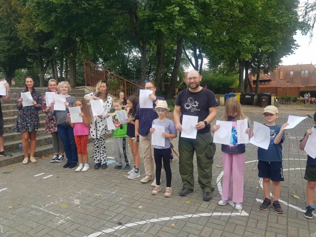 Na terenie, przed szkołą, stoją w szeregu rodzice z dziećmi. Każdy trzyma w ręku podniesioną kartkę. Z liter powstało hasło Bezpieczni na drodze. Za nimi są widoczne schody i drzewa.