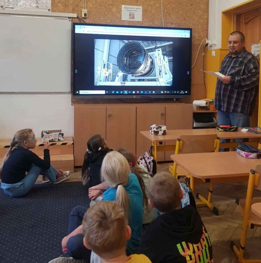 Na zdjęciu widać klasę szkolną. Kilkoro dzieci siedzi na dywanie, tyłem do aparatu. Patrzą na duży ekran, gdzie prezentowane jest zdjecie teleskopu. Obok stoi mężczyzna z kartką w ręku. Po lewej stronie ekranu znajduje się biała tablica. Po prawej stronie stoją stoliki.