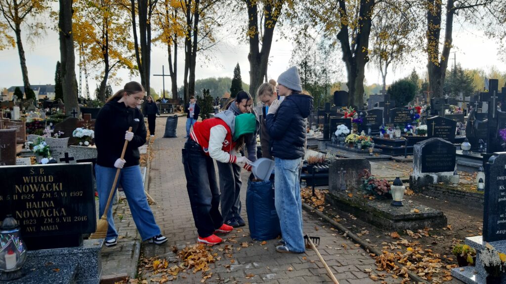 Zdjęcie przedstawia grupę osób stojących na alejce na cmentarzu. Trzymają one worek na śmieci i wkładają do niego liście. Jedna osoba w biało czerwonej bluzie ma zielony kaptur na głowie i pochyla się nad workiem. Obok grupy stoi dziewczyna z miotłą i zamiata liście.