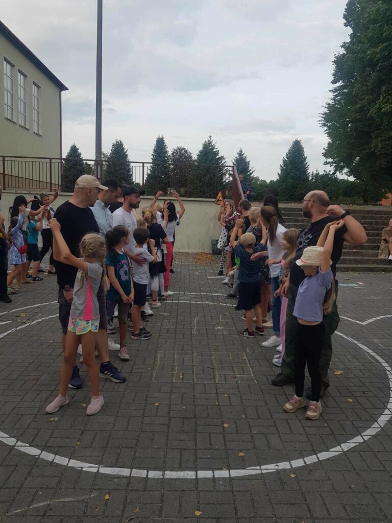 Na zdjęciu widać 3 rzędy osób, składające się z dzieci i rodziców. Zawodnicy podają do siebie znak drogowy. Kilka osób podniosło ręce w górę, na znak zakończenia konkurencji.