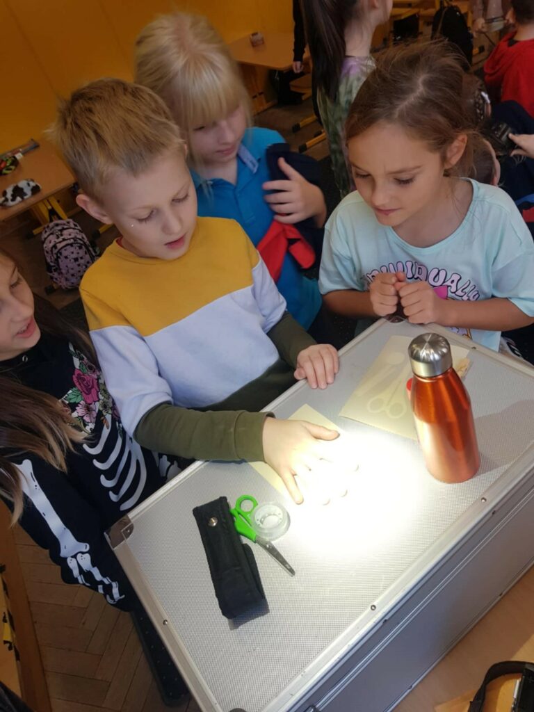 Zdjecie przedstawia salę szkolną. Na ławce leży srebrna walizka. Na niej położony jest arkusz papieru fotograficznego, a na nim są różne przedmioty: nożyczki, taśma klejąca, kartka, stoi bidon. Na kartce chłopiec poożył rękę. Wokół walizki stoją dzieci i przygladają się. Wszystkie leżące przedmioty są oświetlone.