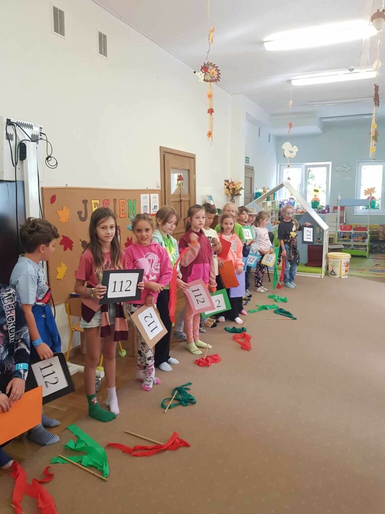 Dzieci ustawione są w szeregu, na długość całej sali przedszkolnej. W rękach trzymają kartki z numerem 112. Przed nimi, na dywanie, leżą szarfy czerwone i zielone. Dzieci są uśmiechnięte, śpiewają piosenkę.