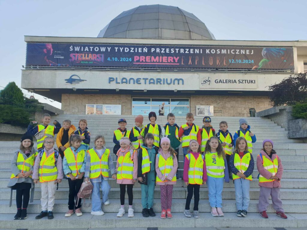 Na zdjęciu widzimy grupę dzieci stojących na schodach przed planetarium. Dzieci ubrane są w żółte odblaskowe kamizelki, co sugeruje, że są na wycieczce szkolnej. W tle znajduje się budynek planetarium z napisem promującym wydarzenie związane z kosmosem, odbywające się w październiku 2024 roku. Baner nad wejściem zapowiada światowy tydzień przestrzeni kosmicznej oraz premiery filmowe. Dzieci wydają się być w dobrych nastrojach, a pogoda jest słoneczna, co sprzyja zwiedzaniu.