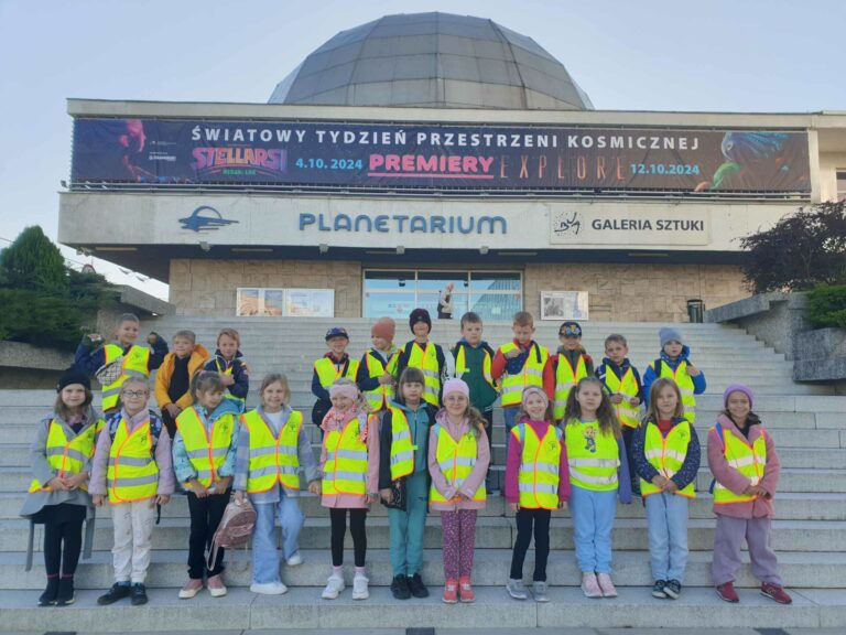Na zdjęciu widzimy grupę dzieci stojących na schodach przed planetarium. Dzieci ubrane są w żółte odblaskowe kamizelki, co sugeruje, że są na wycieczce szkolnej. W tle znajduje się budynek planetarium z napisem promującym wydarzenie związane z kosmosem, odbywające się w październiku 2024 roku. Baner nad wejściem zapowiada światowy tydzień przestrzeni kosmicznej oraz premiery filmowe. Dzieci wydają się być w dobrych nastrojach, a pogoda jest słoneczna, co sprzyja zwiedzaniu.